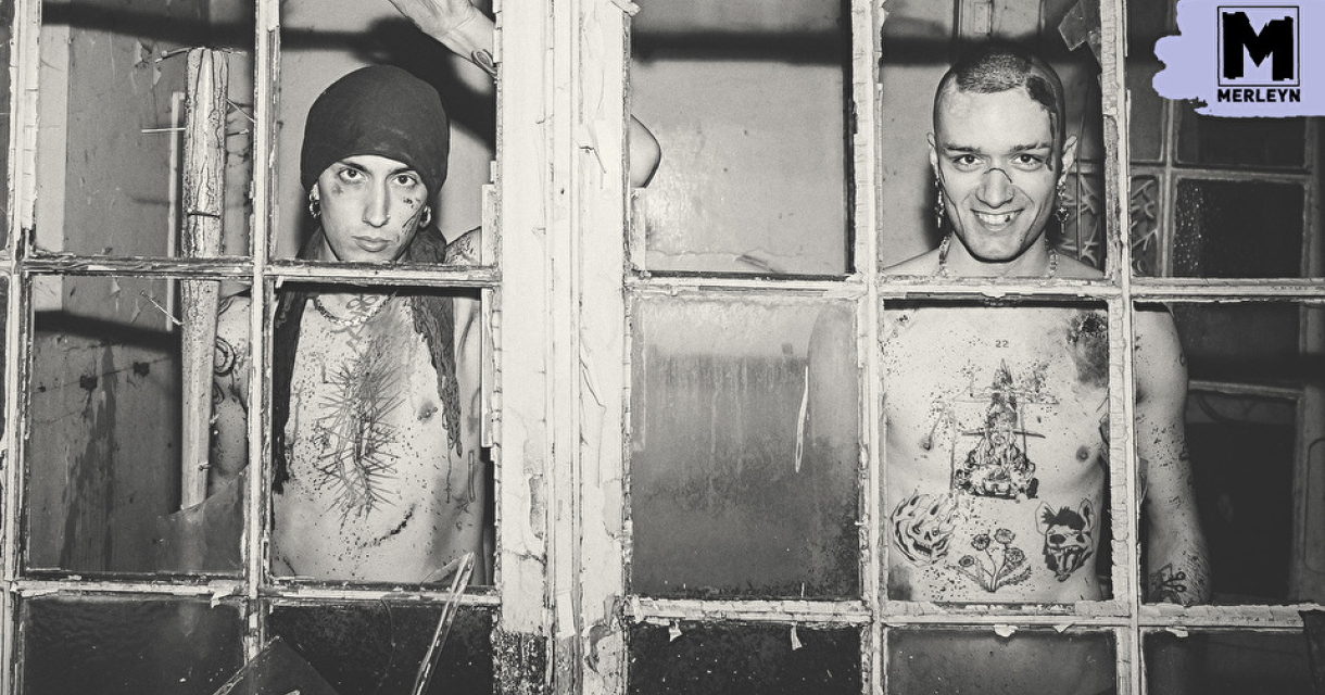 Twee mannen met tattoos staan achter een vergrendeld raam.