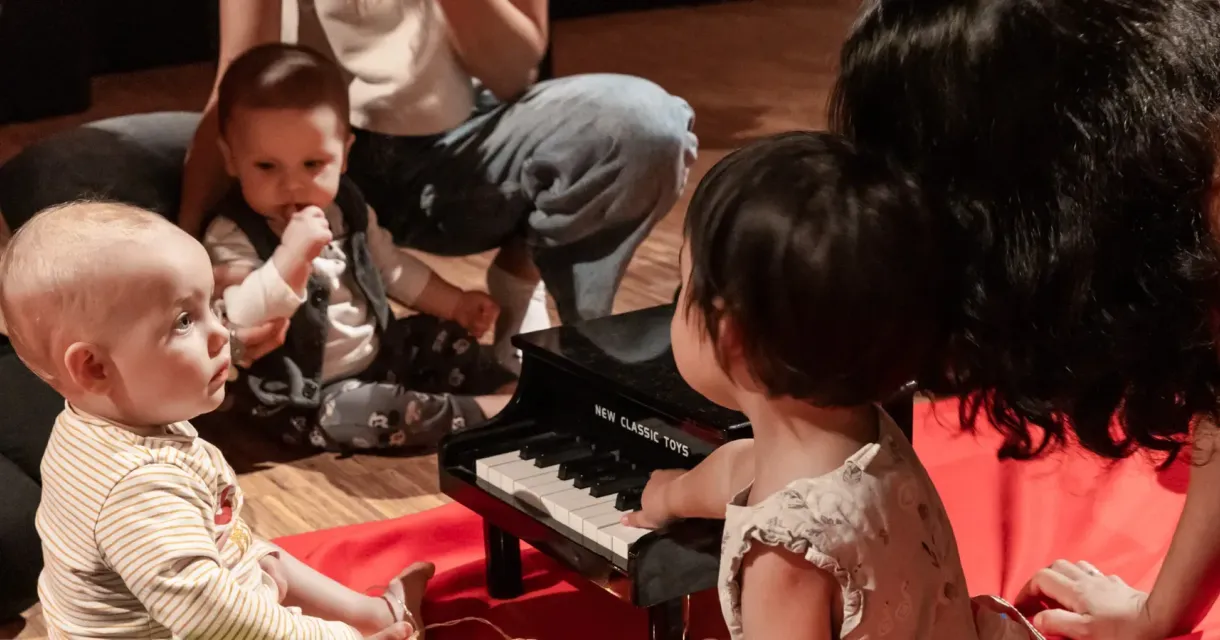 Five children sit on the ground, with two playing together on a mini piano.