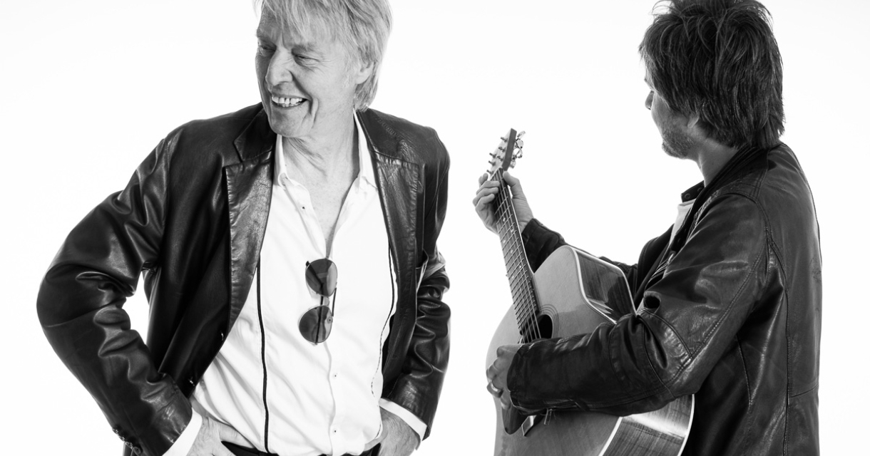 Een lachende man met een zonnebril en een andere man met een gitaar, beiden in leren jassen.