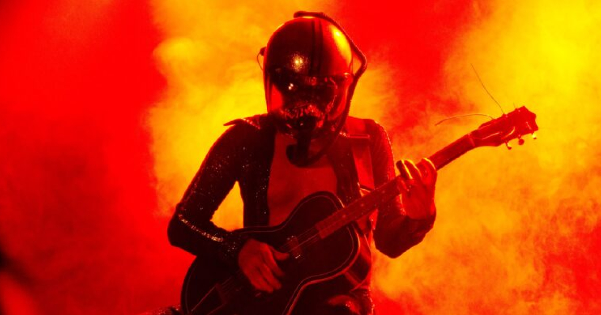 Een muzikant in een ruimtehelm speelt gitaar tegen een achtergrond van felgekleurde rook.