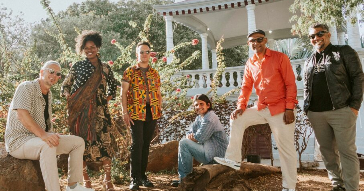 Zes mensen van de muziekgroep CaboCubaJazz poseren in een tuin met bloemen.