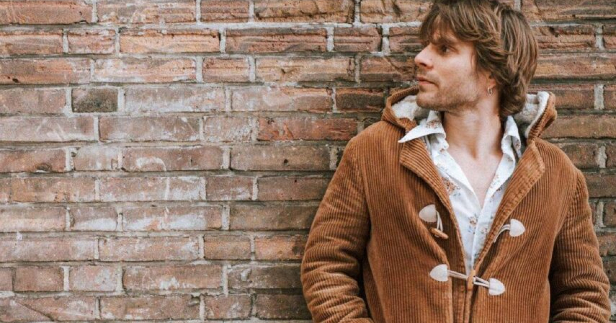 Een man met een bruin vest leunt tegen een muur.