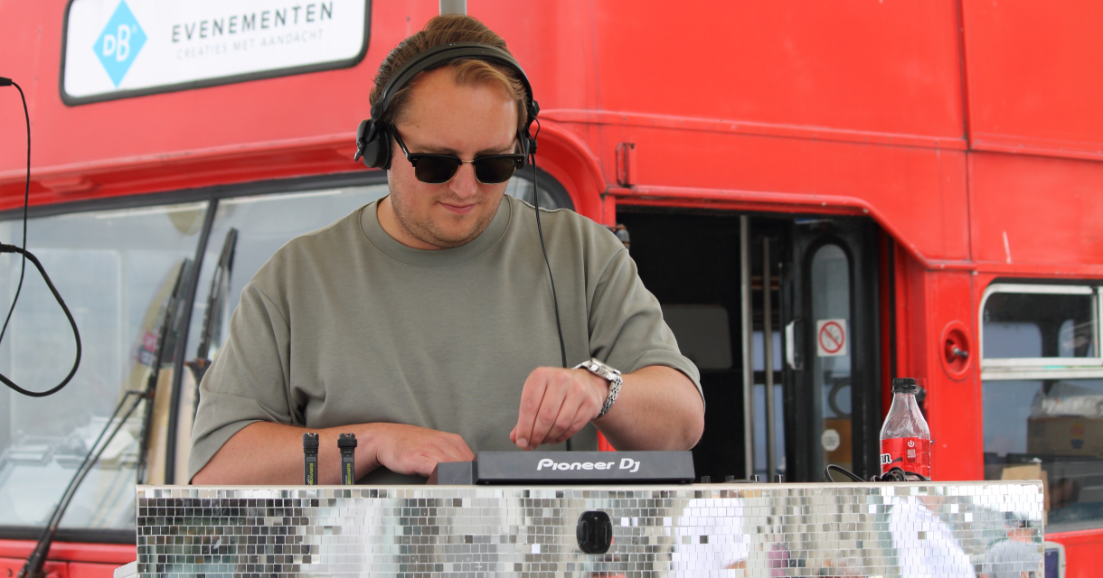 Een DJ aan het draaien met een glanzend mengpaneel, rode bus op de achtergrond.