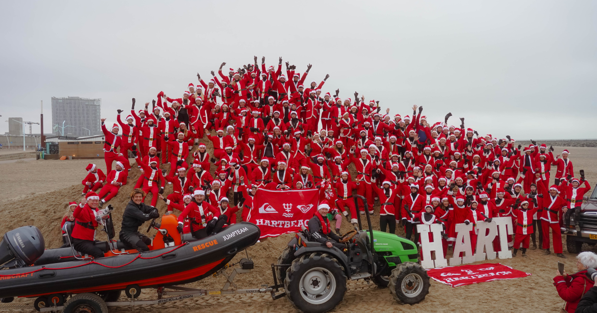 Meer dan honderd mensen in kerstman-outfits op het strand, met een tractor en een boot.