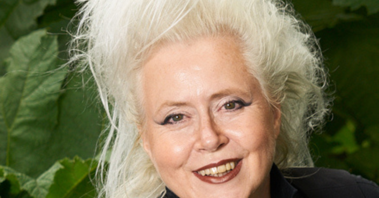 Een vrouw met lang, wit haar en een zwarte outfit glimlacht.