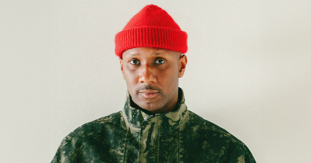 Rapper Dio met rode muts en camouflagejas voor een beige achtergrond, kijkt serieus in de camera; passend bij het concertthema waarin hij zichzelf steeds opnieuw uitvindt.