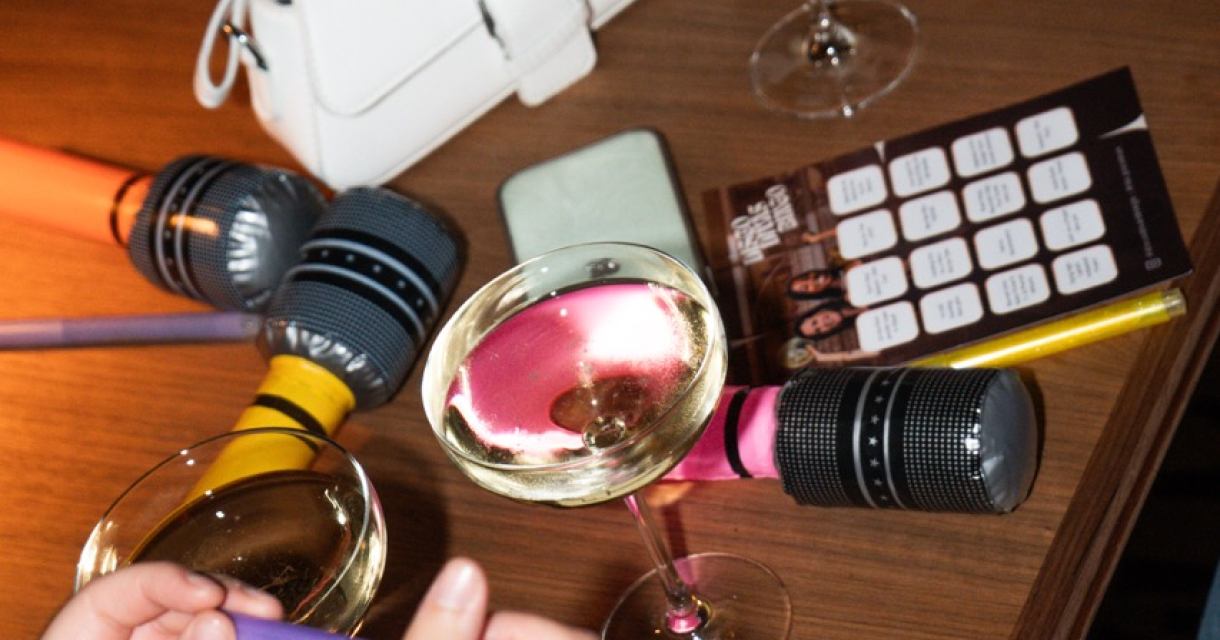 Volle tafel met handtasje, microfoons, glazen champagne, bingoformulieren en handen met stiften.