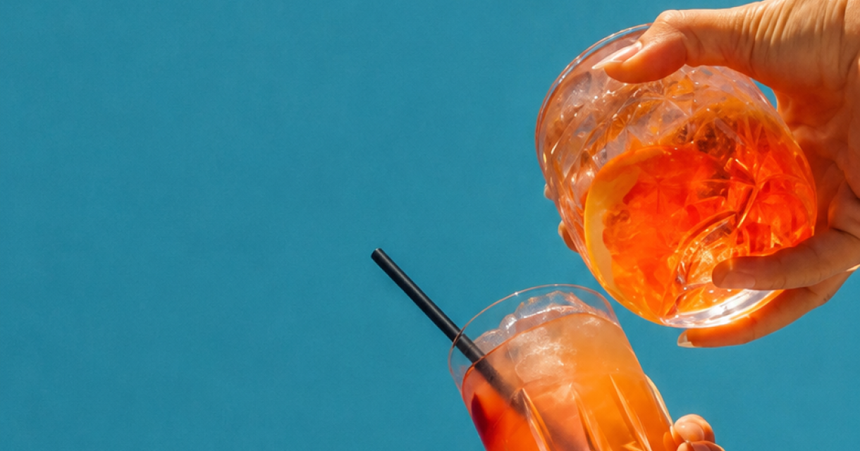 Twee handen houden glazen met oranje drankjes op een blauwe achtergrond.