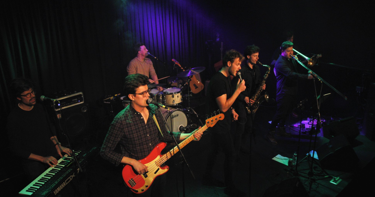 Zes muzikanten spelen live op een podium met instrumenten.