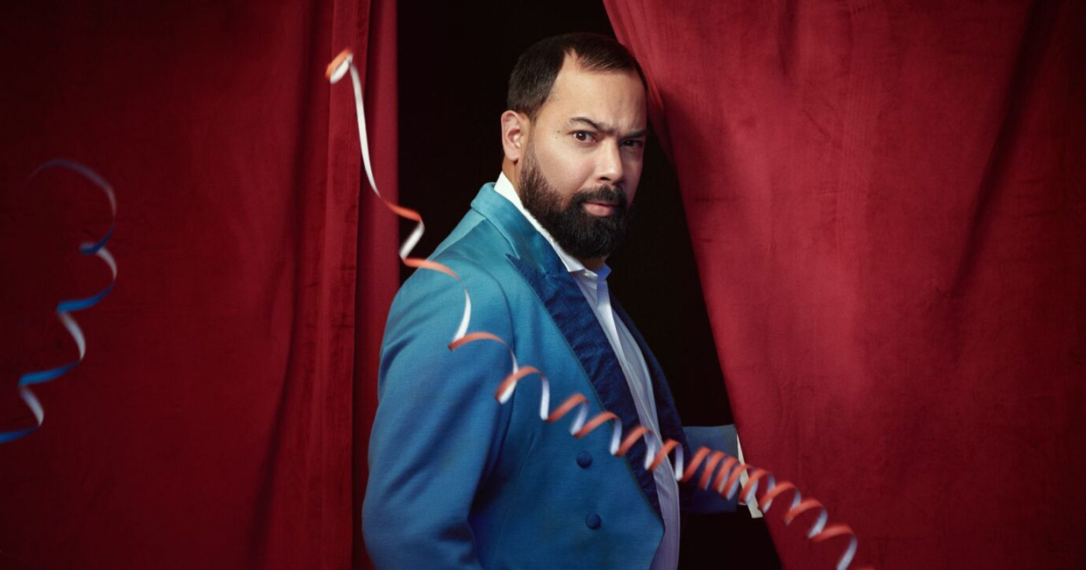 Een man met een baard in een felblauw kostuum staat voor een rode gordijn.