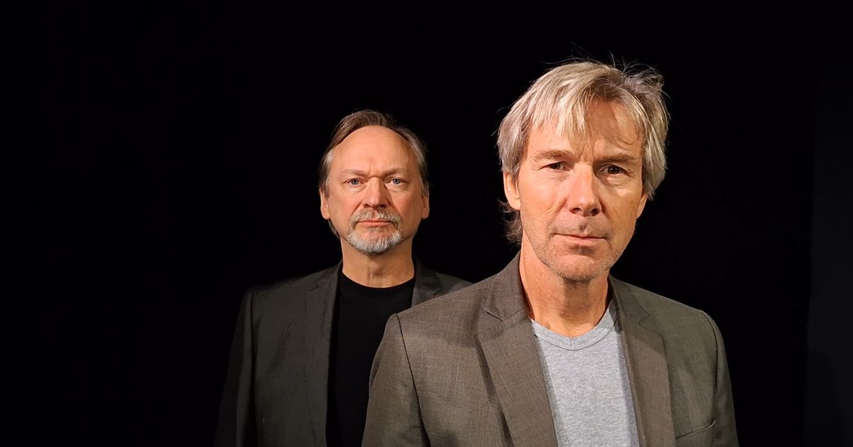 Twee mannen met grijze jassen staan in een donkere ruimte