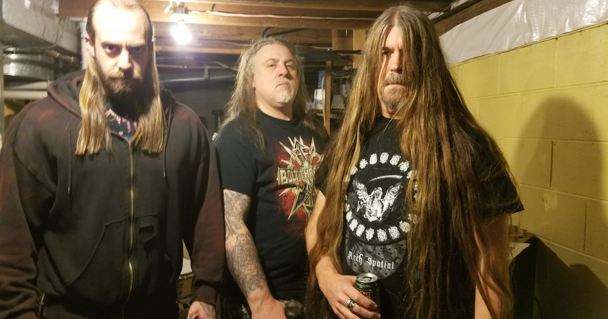 Three members of the band Drawn and Quartered, with long hair and dark clothing, stand in a basement.
