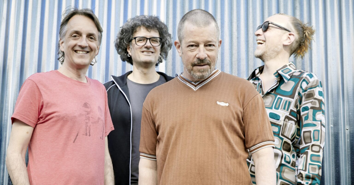 Vier mannen poseren lachend voor een metalen achtergrond.