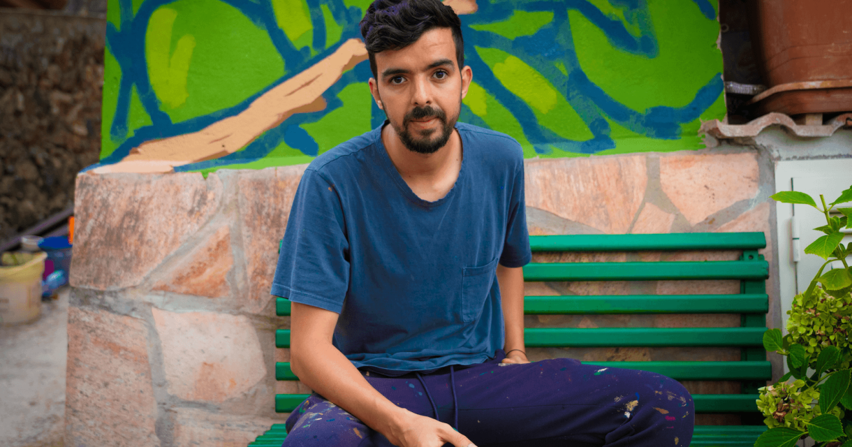 A man sits on a green bench holding a brush, in front of a mural.
