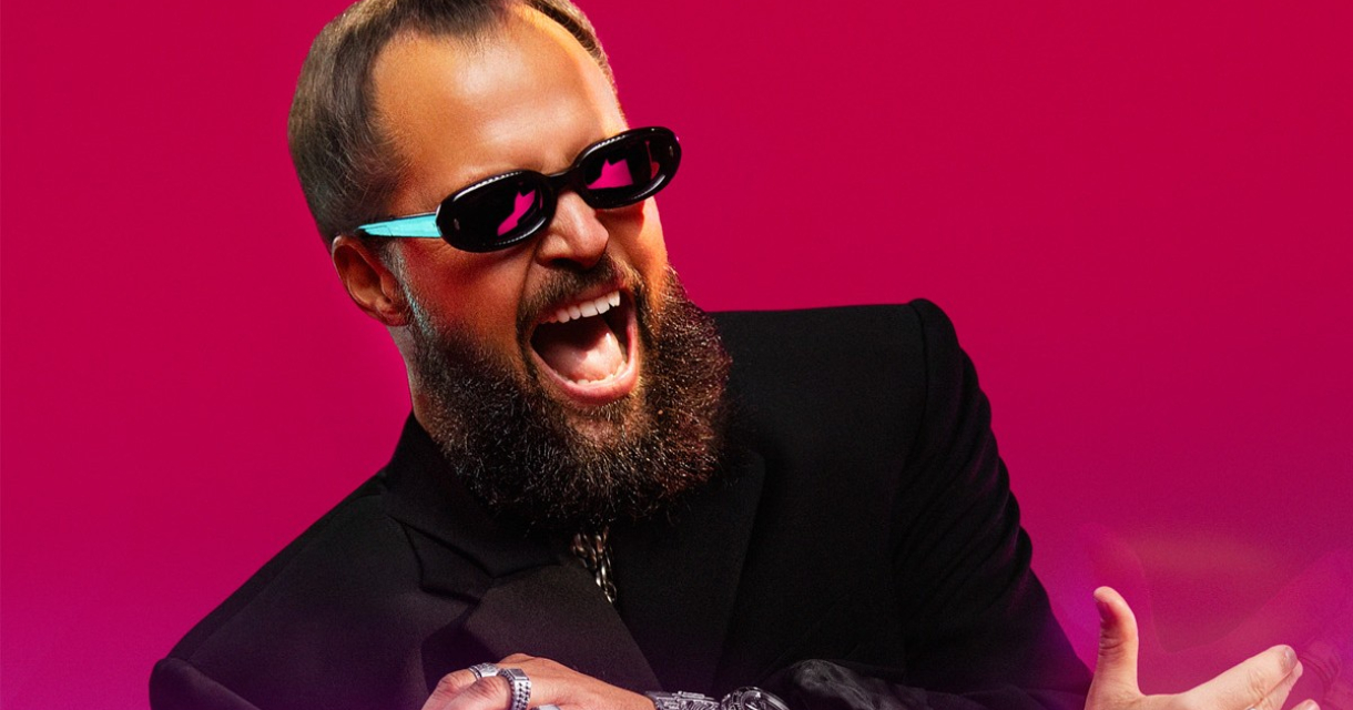 Man met een knot, zonnebril en juwelen, lachend voor een roze achtergrond.