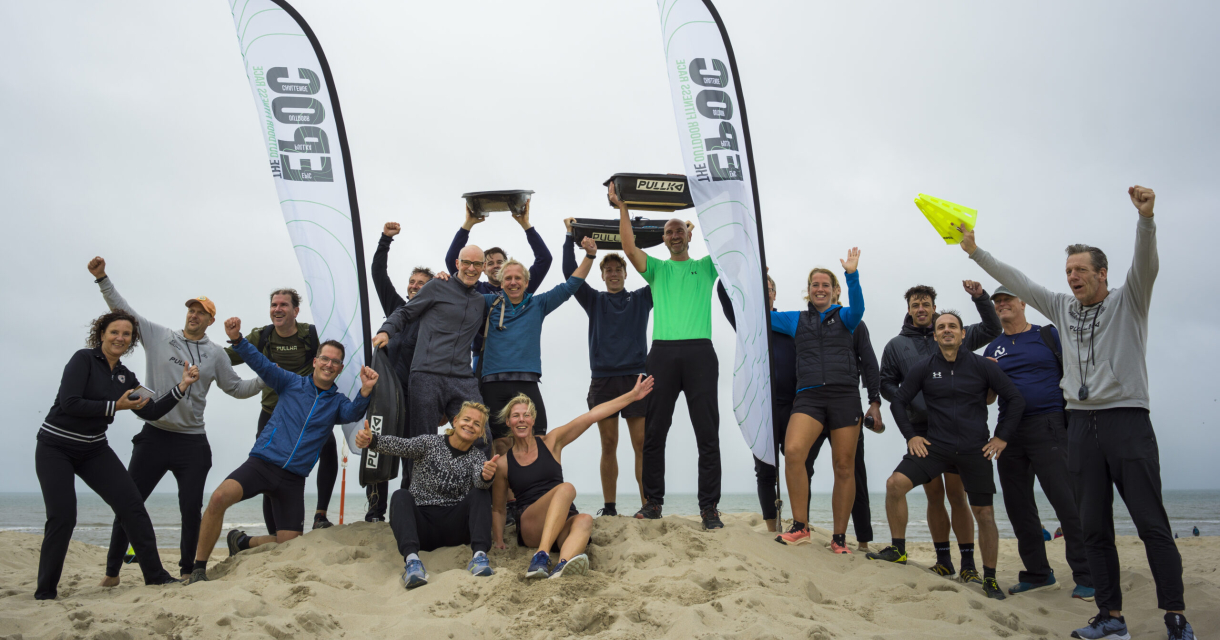 Een groep mensen viert op het strand met trofeeën en vlaggen.