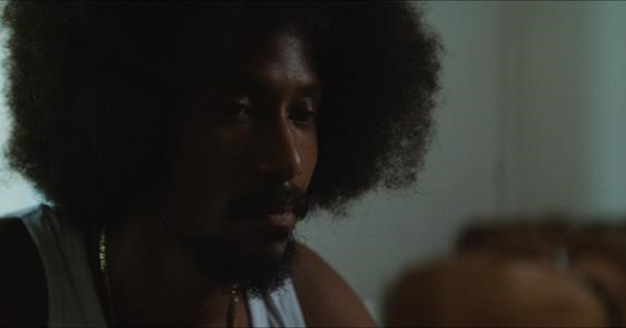 Filmfragment  van een man met afro haar kijkt serieus naar links. Srefidensi Dey – 50 jaar onafhankelijkheid van Suriname