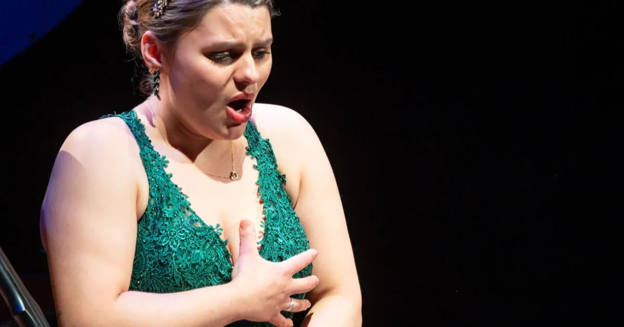 Een vrouw in een groene jurk zingt emotioneel op een podium.