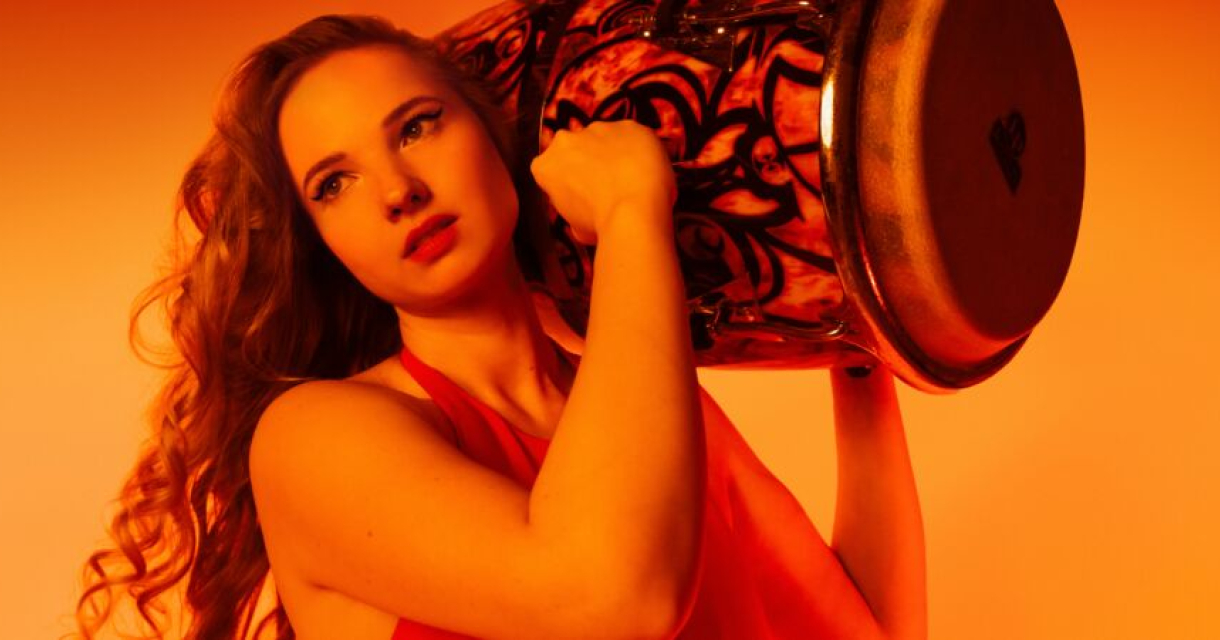 Vrouw met lange krullen houdt een conga vast voor een warme oranje achtergrond.