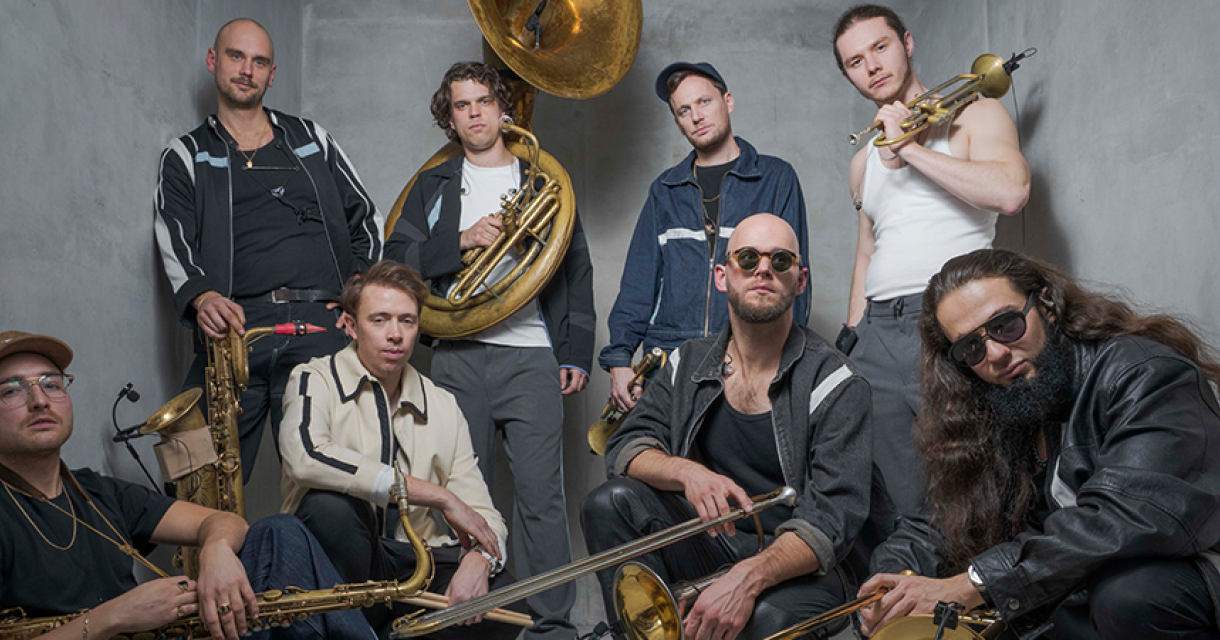 Een groep van acht muzikanten met verschillende instrumenten, inclusief trompetten en saxofoons, in een grijze ruimte.