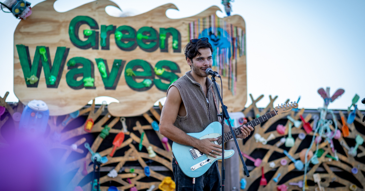 Een zanger met een blauwe gitaar op het podium van het Green Waves festival, omringd door kleurrijke decoraties.