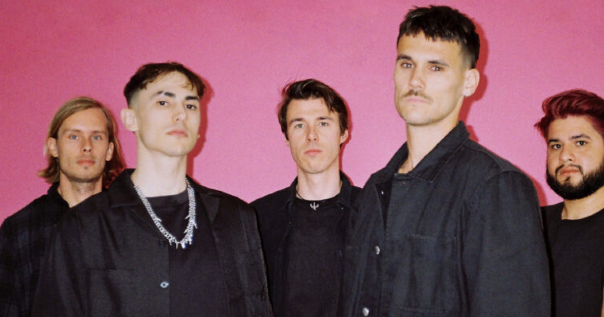 Bandportret voor een roze achtergrond. Vijf leden van band Ann My Dice poseren tegen een roze achtergrond.