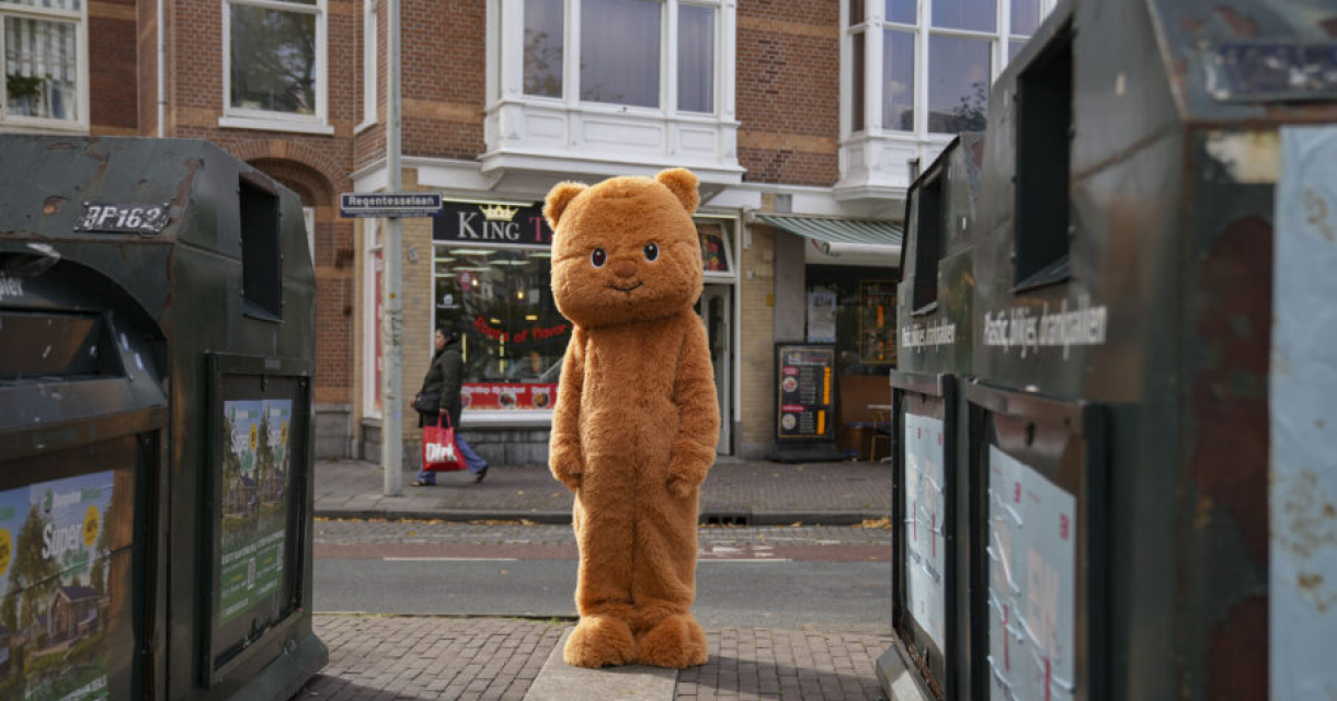 Mascotte in oranje kattenkostuum poseert vrolijk in een theateromgeving, omringd door kinderen en buurtbewoners. De setting ademt een gezellige, laagdrempelige sfeer passend bij een buurtvoorstelling.
