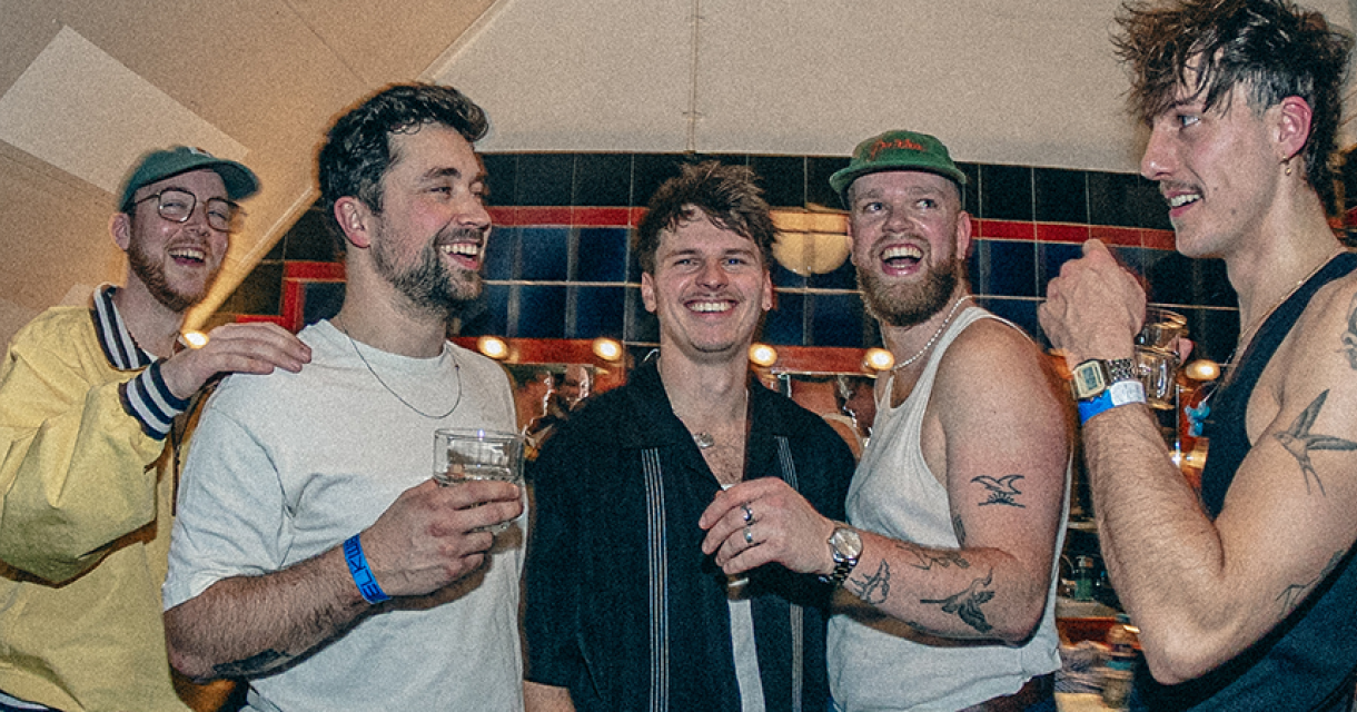 Vijf lachende vrienden met drank in hun handen, gekleed in casual kleding.