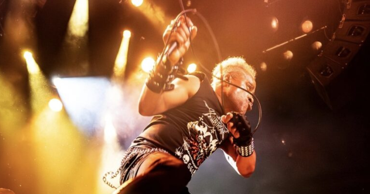 Een performer met blonde haren en een zwart shirt zingt in het licht van een optreden, met een microfoon in de hand.