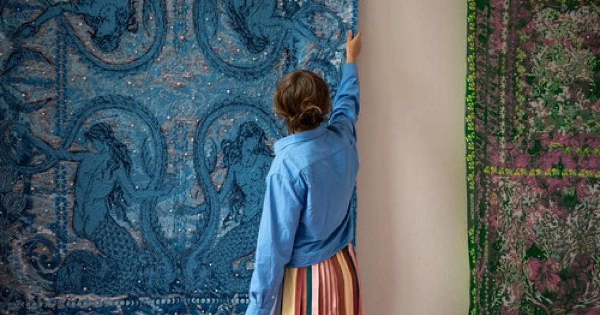 Een vrouw in een blauwe blouse staat voor twee wandtapijten. Foto: Studio Dewi, 2026