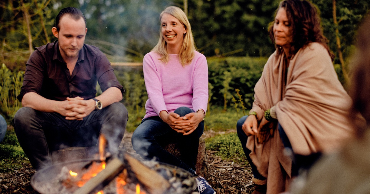 Drei Personen sitzen in der Natur um ein Lagerfeuer.