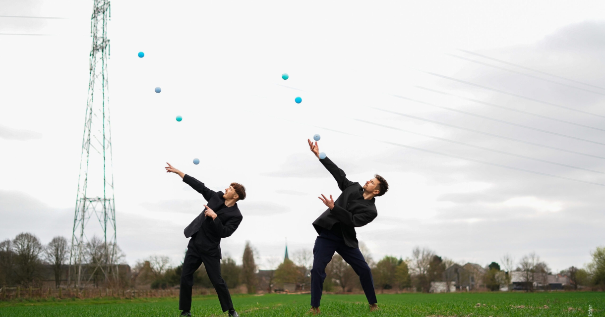 Jam.Shenanigans – Splash. Twee jonge mannen in zwart kostuum jongleren met ballen in een groen veld.