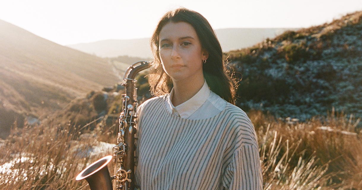 Jasmine Myra met een saxofoon staat in een grazige omgeving met heuvels op de achtergrond.