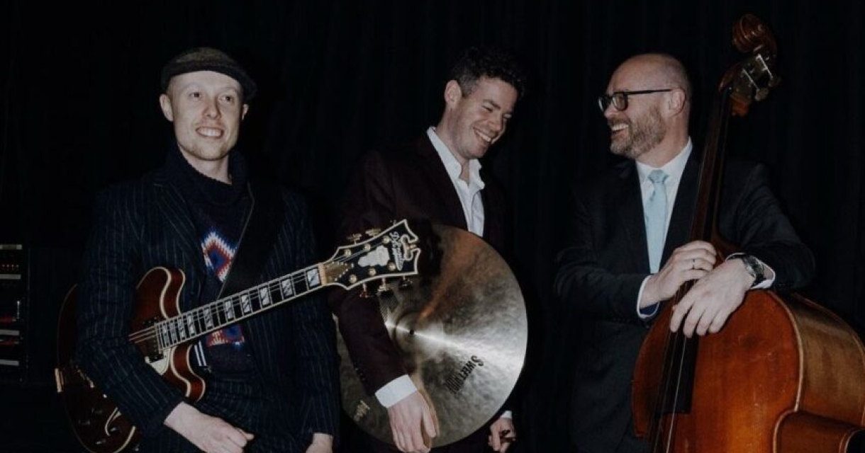 Drie muzikanten met een gitaar, cymbaal en contrabas glimlachen tijdens een optreden.