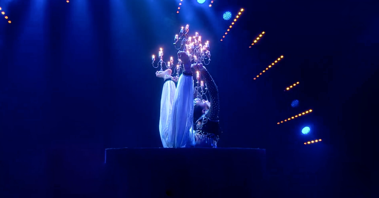 A performer in a dark blue setting with a chandelier, gracefully moving.