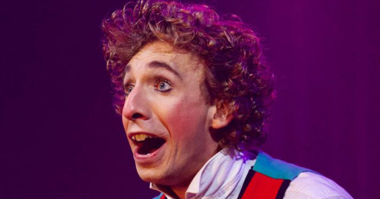 Een jonge man met krullend haar glimlacht breed, gekleed in een kleurrijk kostuum.