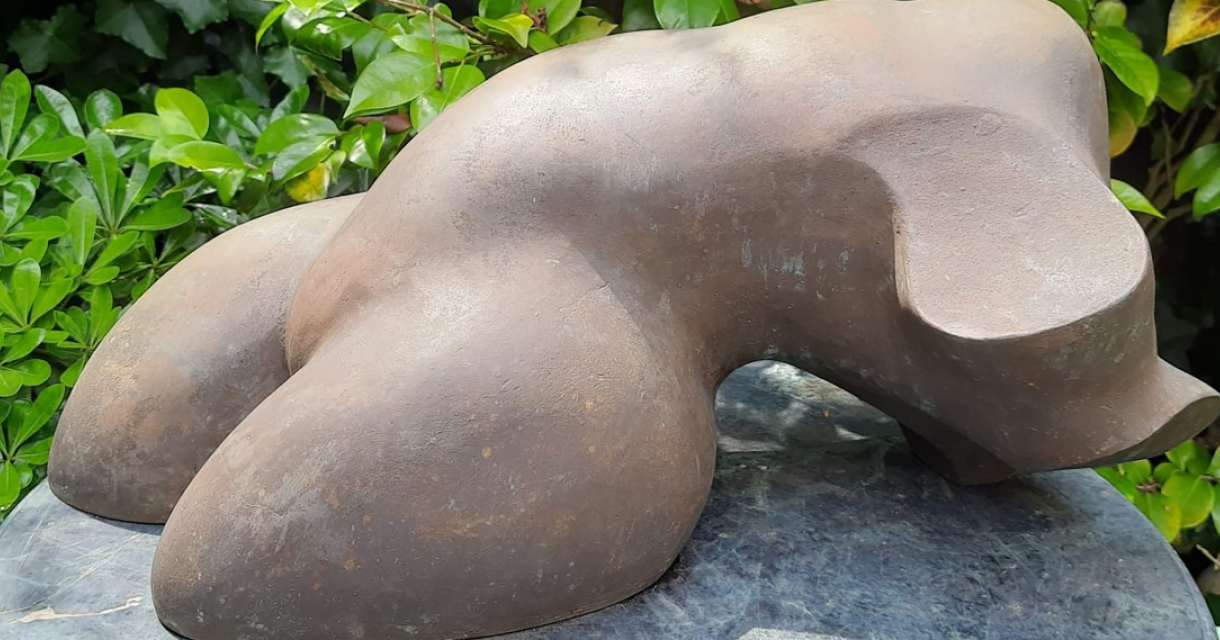 Kim Kroes- Ode aan de vrouw. Abstract bronzen sculptuur met ronde vormen, omringd door groene bladeren.