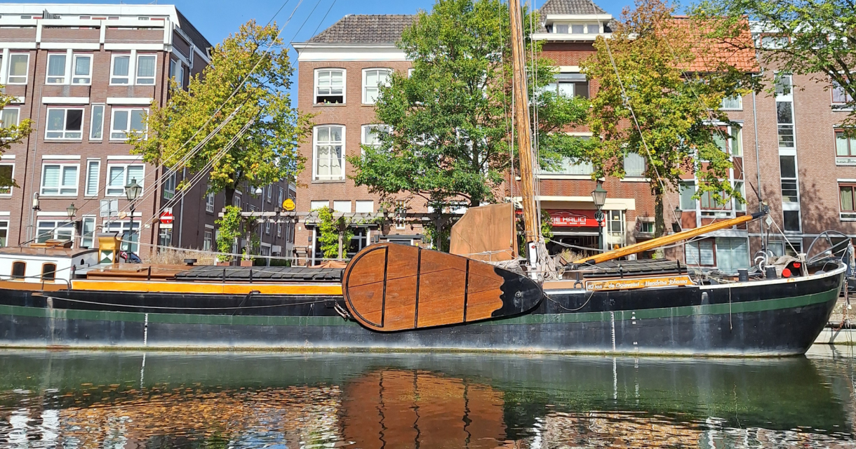 Een historisch schip ligt aangemeerd aan een kanaal in Den Haag, omringd door gebouwen en bomen.