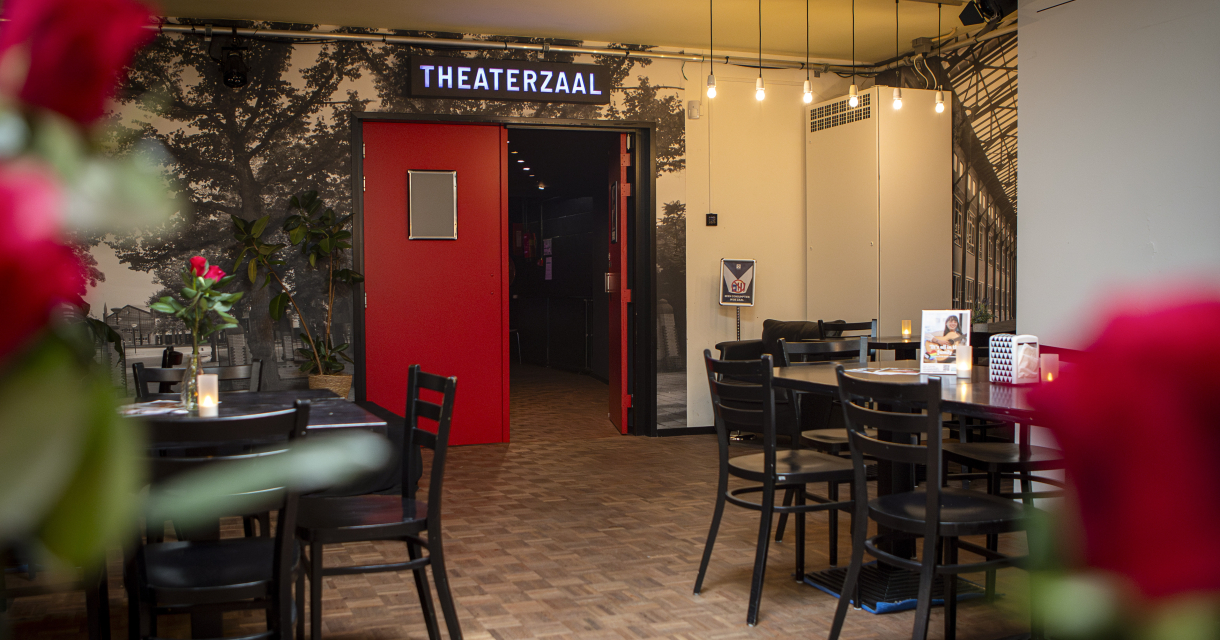 Vue de l'entrée d'une salle de théâtre avec une porte rouge et des tables.