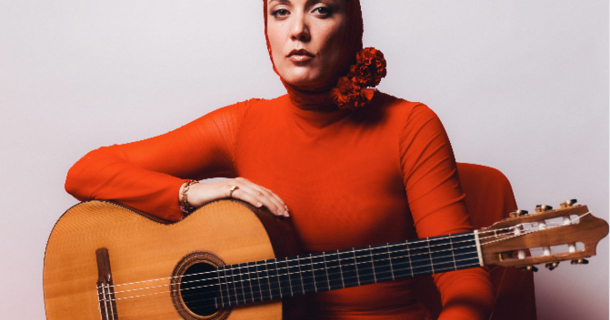 María Marín in een strakke rode jurk met een gitaar, zittend op een stoel.