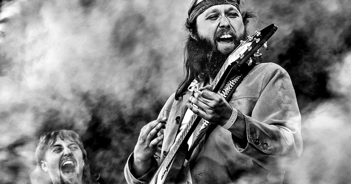 Een rockmuzikant speelt gitaar met veel passie, terwijl een andere muzikant op de achtergrond enthousiast meezingt.