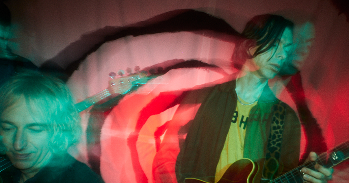 Een band in een kleurrijke, bewegende opname op het podium met muzikanten die gitaar spelen.