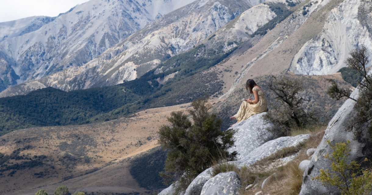 Een vrouw in een gouden jurk zit op een rots met bergen op de achtergrond.