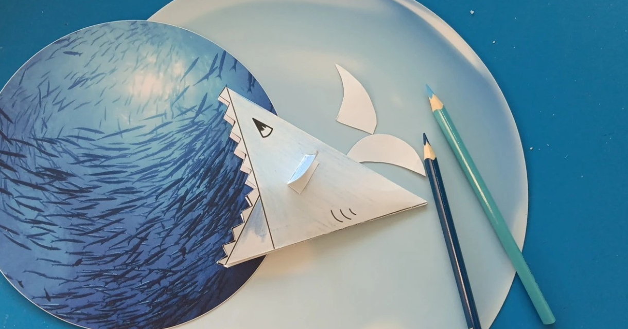 Een papieren haai zwemt in een blauwe oceaan met kleurpotloden.