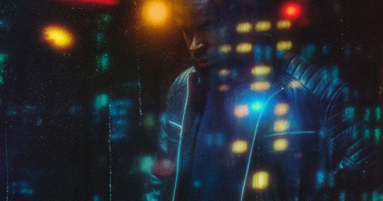 Mansur Brown in een leren jack staat voor een glazen muur met reflecties van stadslichten.