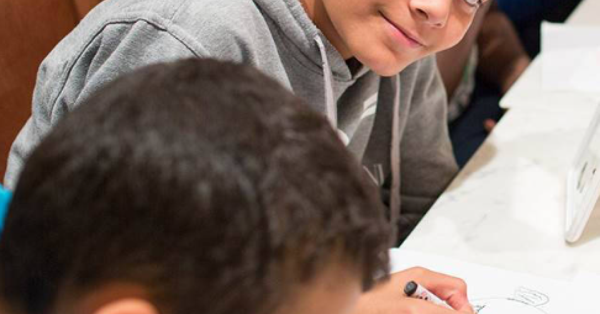 Smiling boy in gray hoodie looks at the camera while drawing.