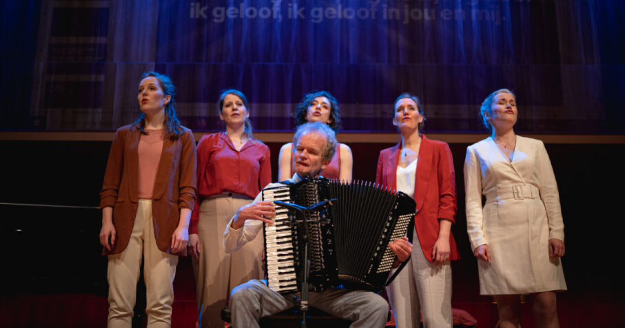 Een man met een accordeon speelt terwijl vijf vrouwen zingen op een podium.