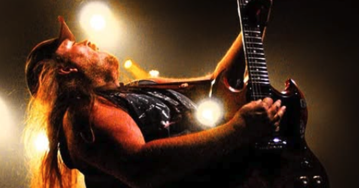 Een man met lang haar speelt electrische gitaar op een podium met lichteffecten.