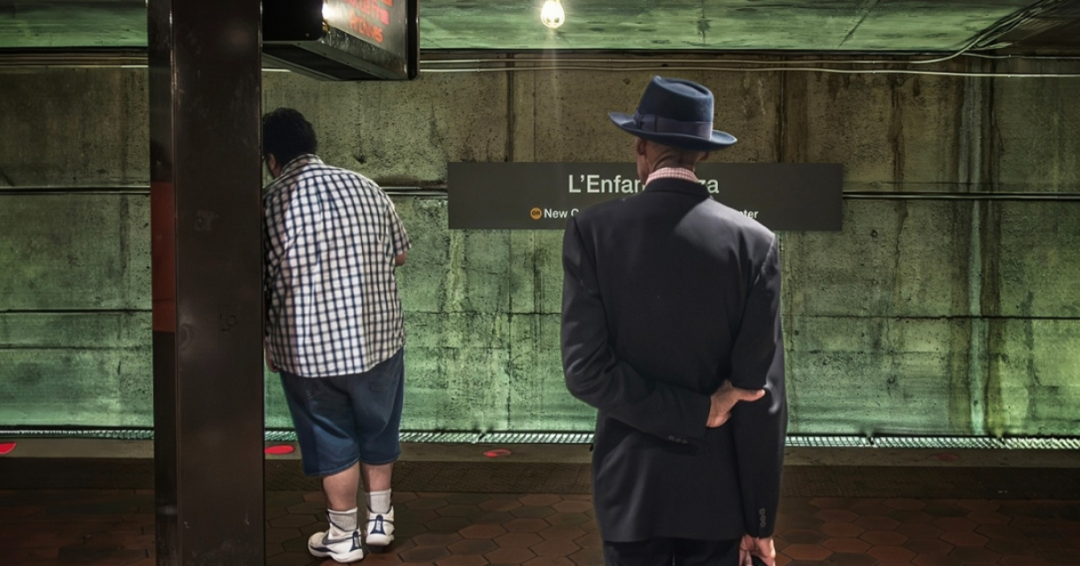 Two men waiting at a subway station, one in casual clothing and the other in a suit with a hat.