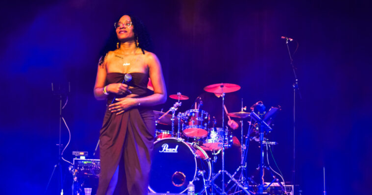 Een vrouw in een lange jurk zingt op het podium met een band achter haar.